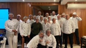 Equipe da Ultracargo participando do Team Building Cooking Show no Experience Lounge, vestindo aventais e chapéus de chef, trabalhando juntos em uma cozinha moderna.